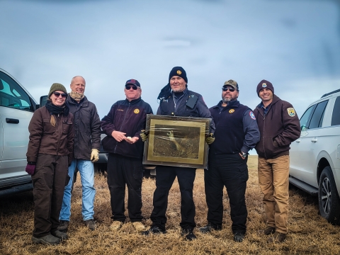 A group of people smiling while the person in the center holds wall art of a black-footed ferret