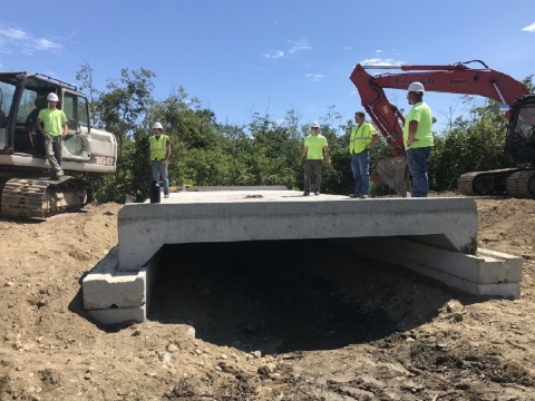 a fresh concrete slab lies across the ditch underneath it. Construction workers stand on top of the large slab and construction vehicles sit in the background. 