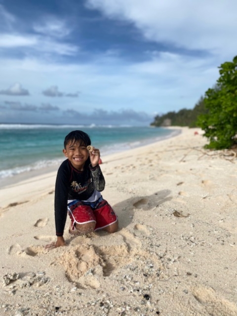 A young boy wearing a black shirt and red shorts sits on a beach holding a shell. The ocean sits in the background.