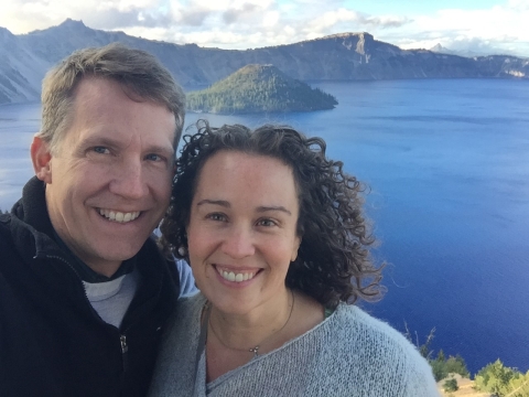 A man and woman smiling in front of a dark blue lake and mountains