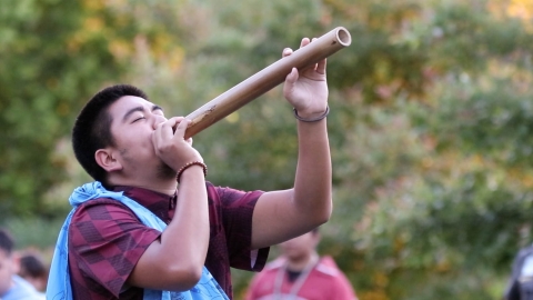 Indigenous person playing a wooden instrument or flute, outdoors. 