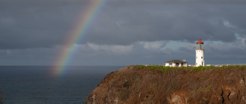 A rainbow arcs over the ocean and a lighthouse sitting on a rocky point. 