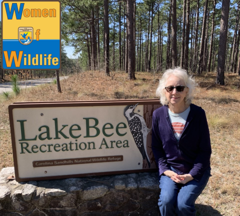 Deb Adams sits on the welcome sign at Lake Bee Recreation area at Carolina Sandhills National Wildlife Refuge. The sign has a woodpecker carved into it. Deb is wearing sunglasses and smiling. The WoW: Woman of Wildlife logo is in the upper left corner. 