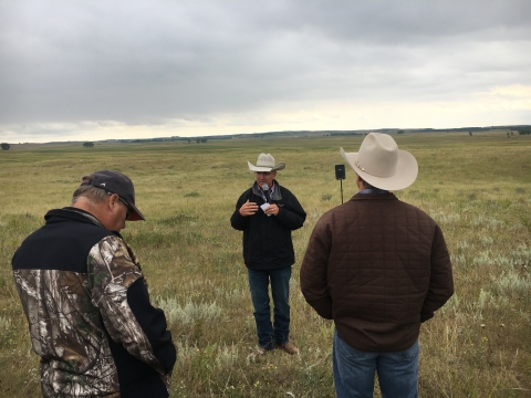 A man wearing a cowboy hat stands in front of a field, speaking to a small crowd of people