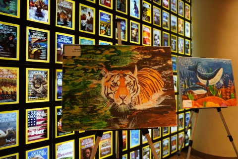 Three examples of artwork on easels and in front of a light panel of magazine covers. 