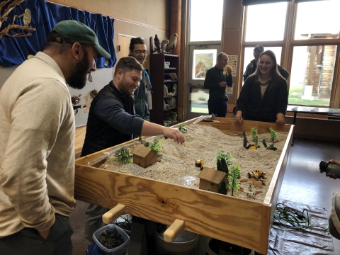 People standing around a table filled with gravel, miniature trees, and miniature sheds