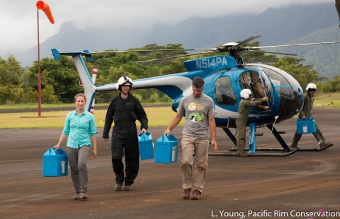 Members carry blue boxes across a helipad with a blue helicopter in the background.