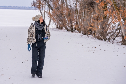 A woman wearing a warm jacket, hat, gloves, and binoculars walks on a snow covered path looking into the trees.