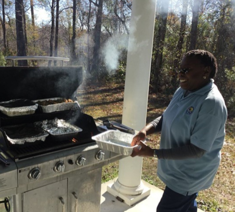 Brenda Williams, volunteer at Waccamaw National Wildlife Refuge, smiles as she holds a tray and stands near a grill where food is being cooked.