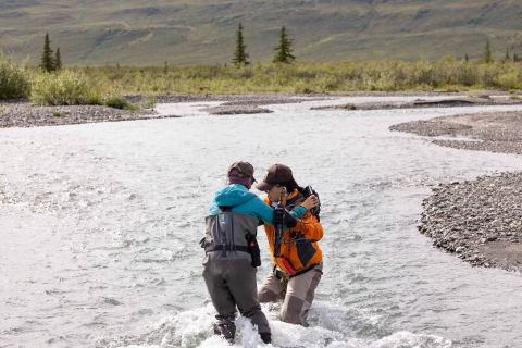 Two people performing a water crossing in a fast flowing river with water reaching their knees with a tundra landscape in the background. 