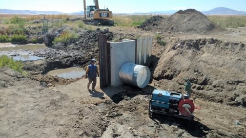 A man in a welding helmet working outdoors with heavy machinery in the distance