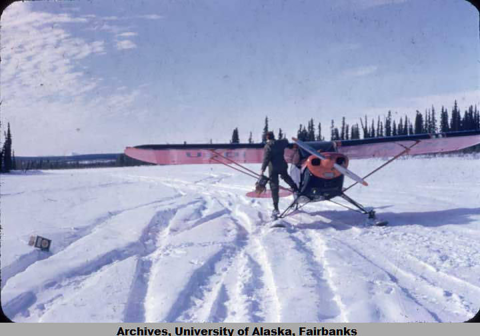 Photograph shows a man hanging off of a Fish and Wildlife Service airplane. He appears to be holding a fuel filter in his hand. 