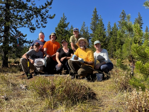 A group of people kneel smiling in an alpine meadow. One of them holds a bumble bee nest on a paper plate.