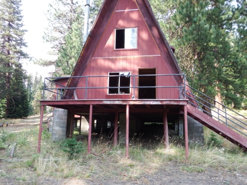 A red a-frame house on stilts with stairs sits in a wooded fielded