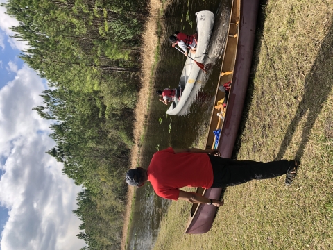 A person watches as two people paddle away in a canoe. There is another canoe parked on shore.