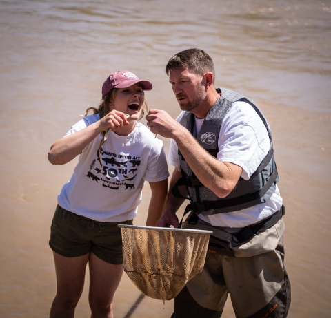 Two people holding a net and kissing some razorback sucker while laughing