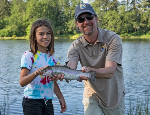 Male Service employee stands beside Big Brothers Big Sisters event participant as he holds a rainbow trout she caught. Lake in background. 