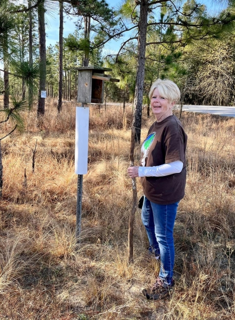 A woman in jeans and long sleeves stands near a bluebird box