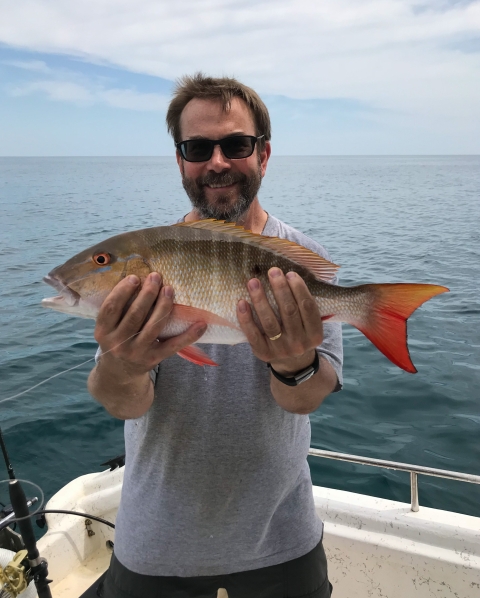 a man wearing sunglasses stands in a boat while holding a gray and orange fish up to the camera. Behind him are water and sky with some light clouds.