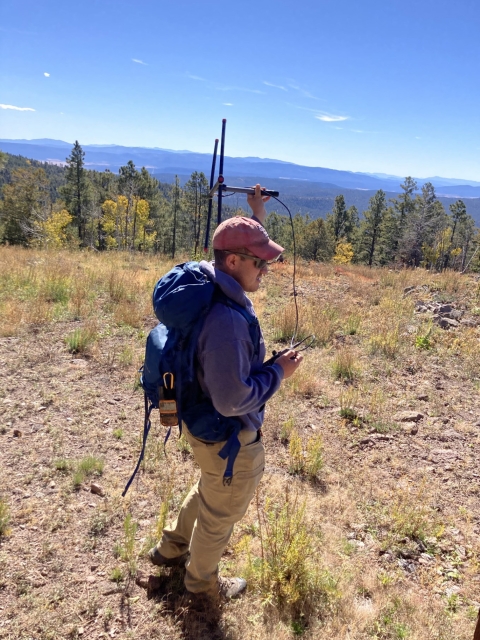 A man stands in a clearing in the forest, holding a radio receiver in the air.