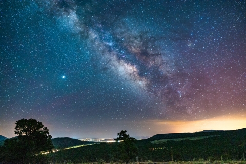 A bright light obstructs the night sky and milkyway directly over a city at night with hills and trees in the foreground and stars pictured in the background.