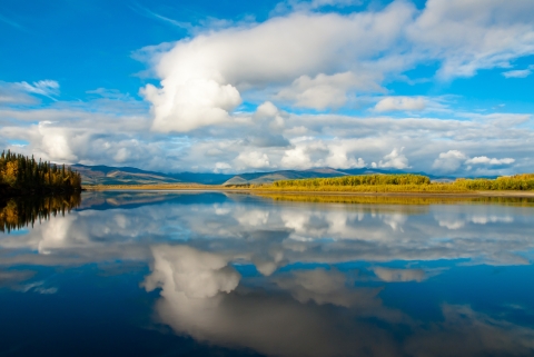 Still water providing a mirror image of the clouds and blue sky above with vegetation on both left and right banks of the water with mountains on the horizon. 