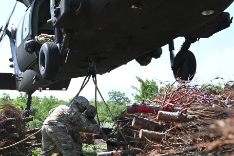 Soldiers in uniform attach stumps of trees to helicopter hovering above