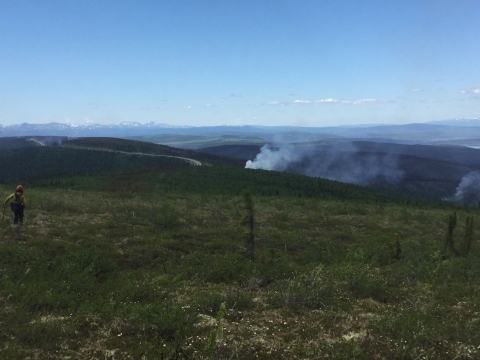 Smoke rises in from distant hills. A wild fire fighter looks across the valley.