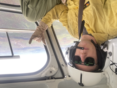 Eric Johnson give a hang lose sign in the helicopter as it flies over burned landscape.