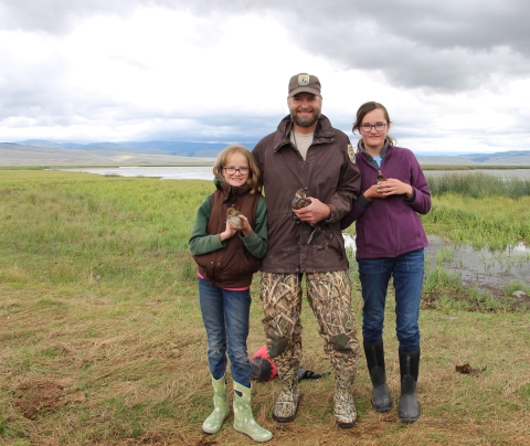 A biologist and children stand together next to a lake holding ducks.