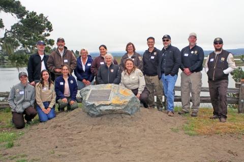 14 people pose behind boulder with a plaque -wooden railing and body of water in the background