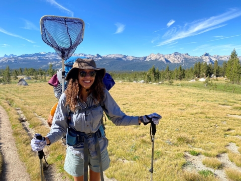 A woman smiles for the camera while holding hiking poles and while wearing a backpack with an attached net on her back