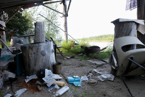 A color photo of Chief Rhonda Pitka's grandmother's fish camp, which has been abandoned for four years due to a lack of salmon on the Yukon River. Buildings are weather-beaten, some laying across the grass while small belongings are strewn on the ground. Boards and metal sheets lay askew beneath the deep green trees.