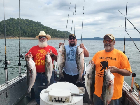 Three men smiling holding a striper in each hand on a boat on a lake 