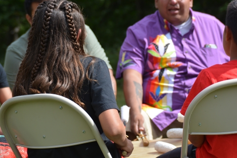 People sitting around a Tribal Ceremonial drum, hitting the top with fur-tipped drumsticks.