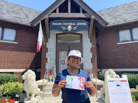 a person holds up a piece of paper in front of a building with a sign that reads "Italian Cultural Center"