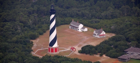 Aerial view of a lighthouse, with a black-and-white barber-striped tower, surrounded by woods.