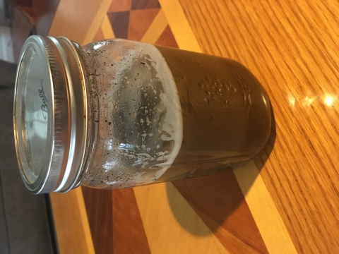 A Mason jar with a lid containing brown liquid with white foam sits on a wood-inlay table