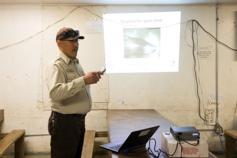 Chris Tulik giving a presentation with a projector on a blank wall holding a controller for the slide show. 