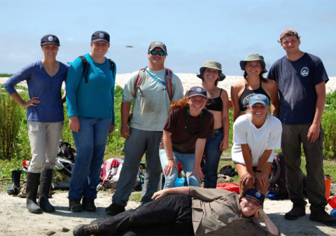 A group of young men and women smiling in front of a white sandy hill. One person poses lying down in front of the group.