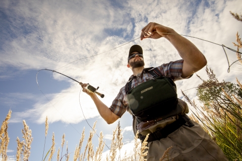 A man fly fishes under a cloudy blue sky.