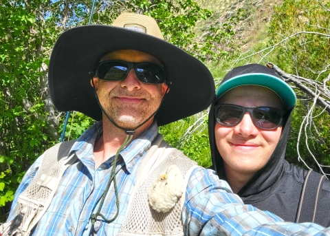 A close up image of two men in fishing gear surrounded by green trees.