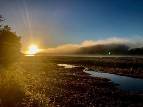 The rising sun, low on the horizon, lights up a river and river plain in the foreground.