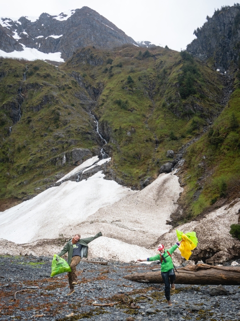 Staff happily carrying trash bags from clean rocky beach with snow covered mountains in background.