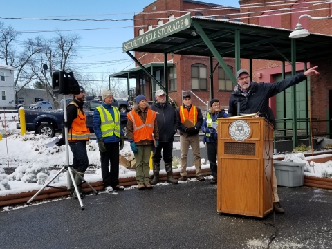 A man in a black jacket and ball cap speaks from behind an outdoor podium with six people standing to his right and a brick factory building behind him.