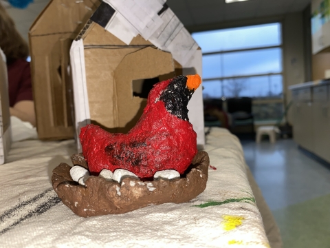 Close up of red male cardinal bird made out of paper clay and sitting in a brown nest with white eggs. In the background is a cardboard bird house and a view out floor to ceiling windows at a gray blue sky.