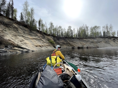 Tim Ericson, sitting at the front of a boat, paddles on the Deshka River. The photo is taken from the back of the boat, and Ericson looks back at the camera over his shoulder. The boat is filled with supplies, including moose antlers. 