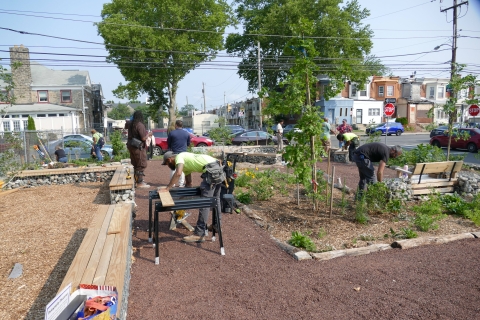 People work on various construction tasks in a gravel lot with city blocks in the background