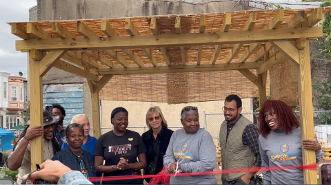 People standing side by side facing the camera underneath a trellis behind a ribbon tied to the trellis and suspended at waist height. One person is holding scissors.