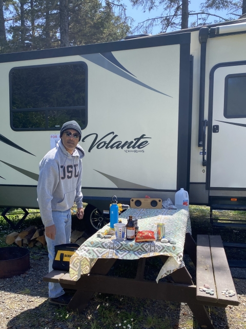 A man wearing a black beanie and sunglasses stands in front of a picnic table and an RV in a camp site. Blue skies and green pines are in the background. 